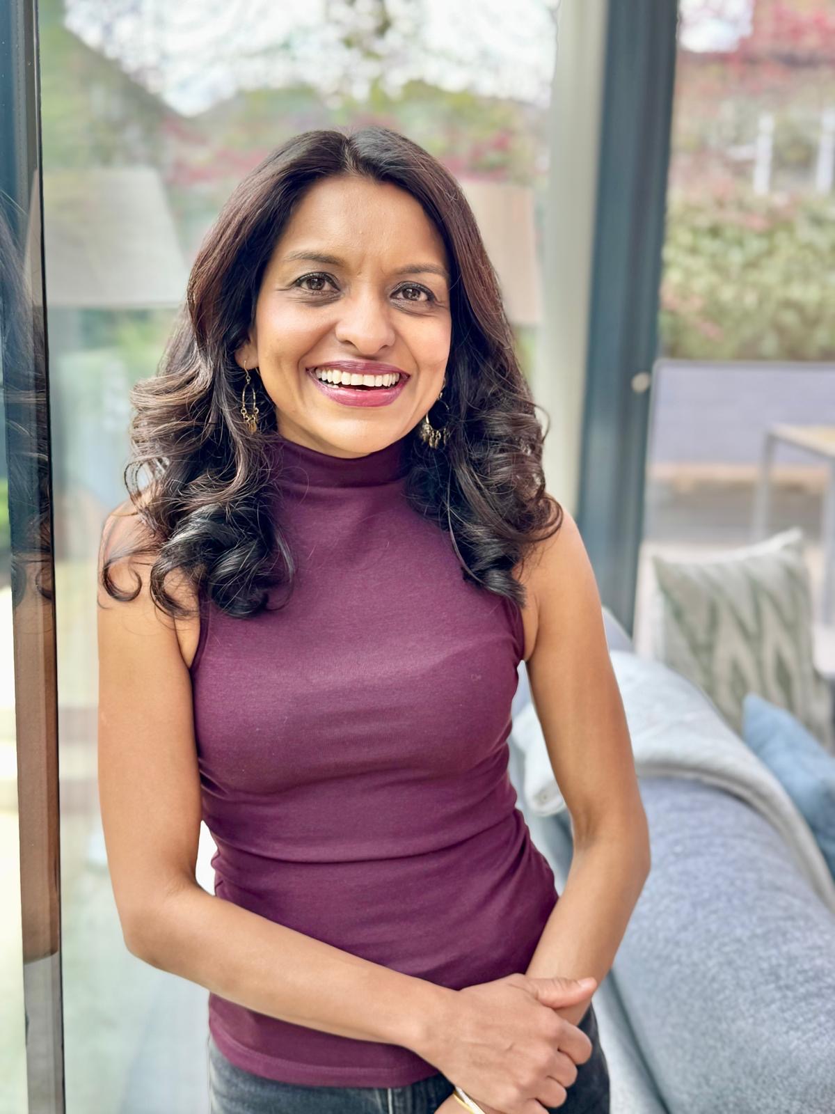 Sabina smiling beside a window in a sleeveless burgundy top