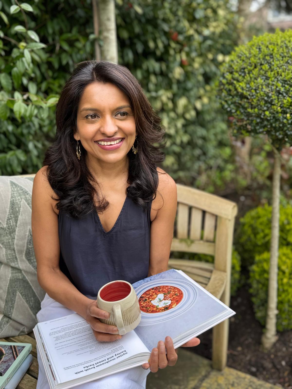 Sabina smiling with a cookbook and a mug in the garden
