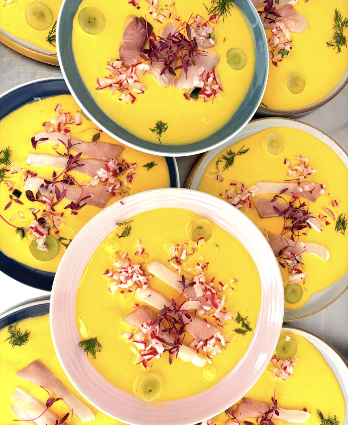 Bowls of a golden chilled course topped with fish, radish, grapes and micro herbs, laid out for a private dinner
